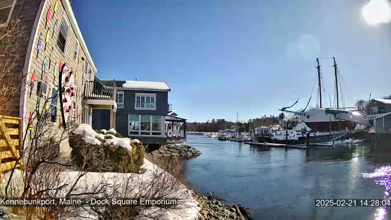 A waterfront view on a sunny day shows snow-covered ground and buildings near a calm river with a large sailboat docked among other smaller boats.