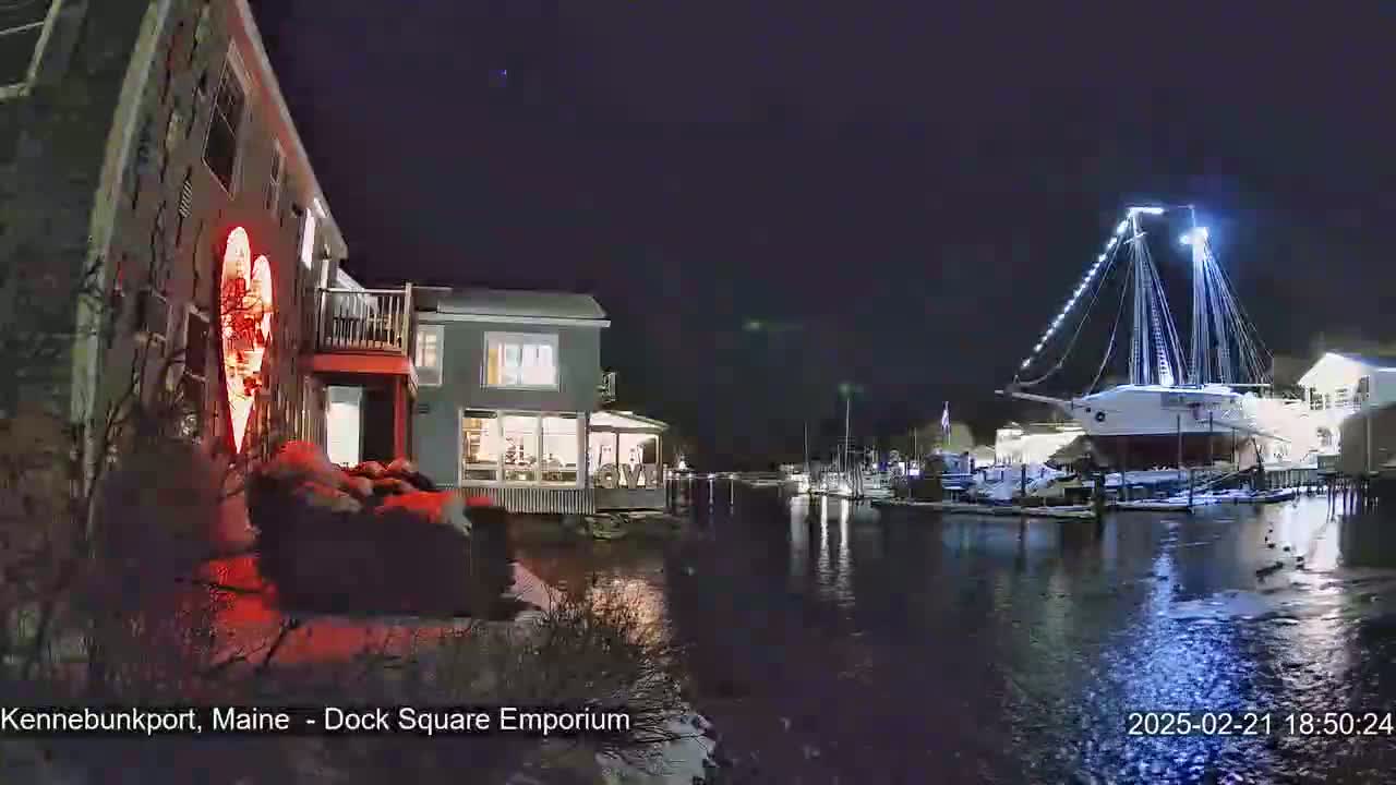 A nighttime waterfront scene shows a building with a large illuminated heart-shaped decoration next to a canal, across which a large illuminated sailboat is moored.