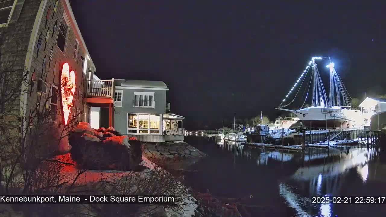 A nighttime view of a harbor with a large sailboat illuminated with white lights, buildings with exterior lights, and snow on the ground.