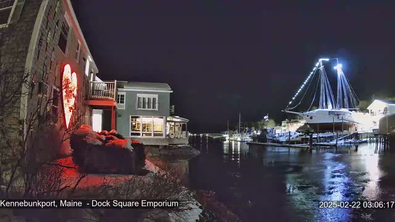 A nighttime scene shows a waterfront with a building displaying a large illuminated heart, other buildings, and a large sailboat docked, all under a clear, dark sky with some snow on the ground.