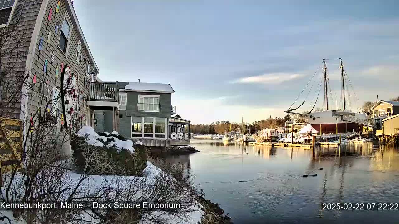 A snow-covered waterfront scene shows buildings, a large sailboat, and a calm river under a partly cloudy, bright sky.
