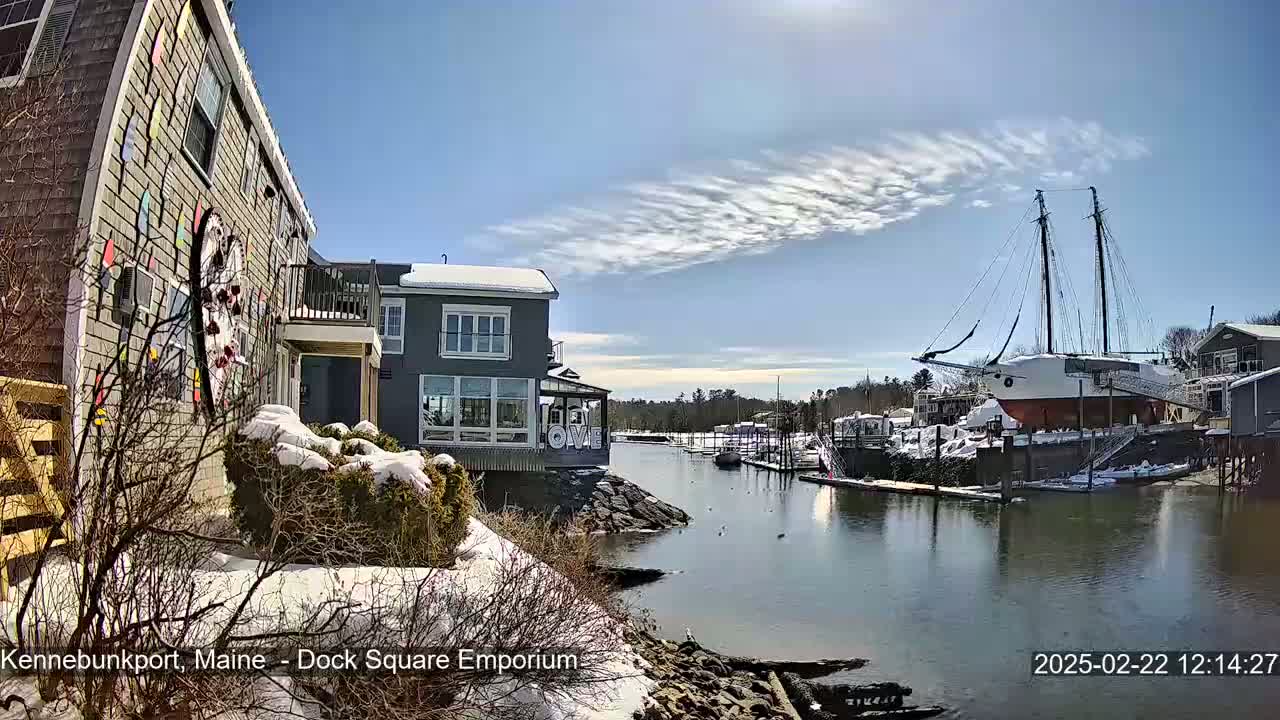 A snow-covered waterfront scene features colorful buildings, a large sailing ship docked, and a partly sunny sky with wispy clouds.