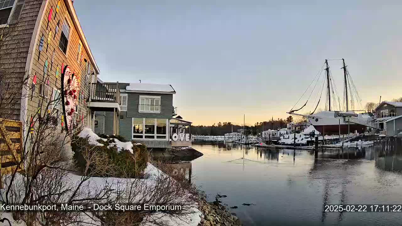 A snow-covered waterfront scene shows a building with colorful nautical decorations next to a calm river with a large sailboat docked, under a twilight sky.
