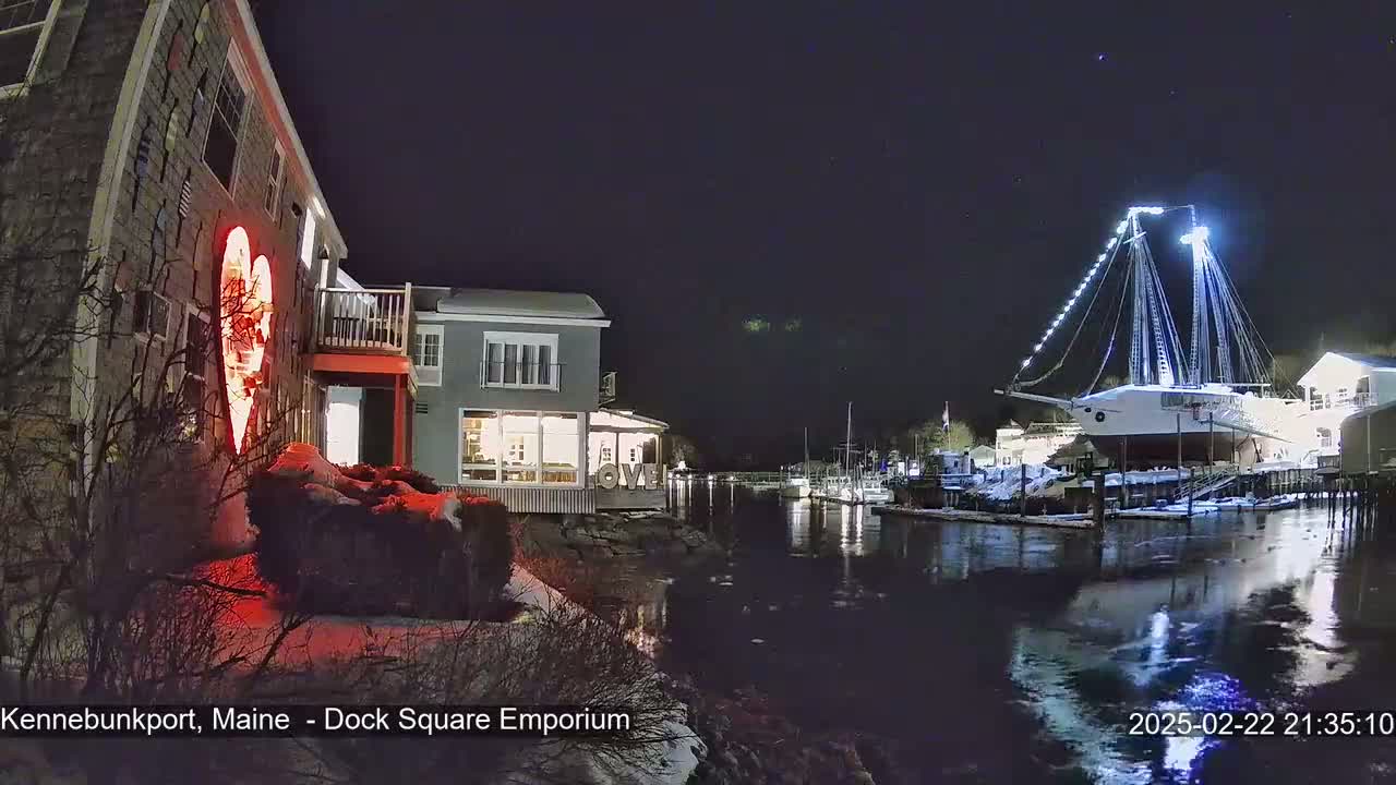 A nighttime waterfront scene shows a building with a large illuminated heart, other buildings, a large illuminated ship, and a calm body of water with snow on the ground.