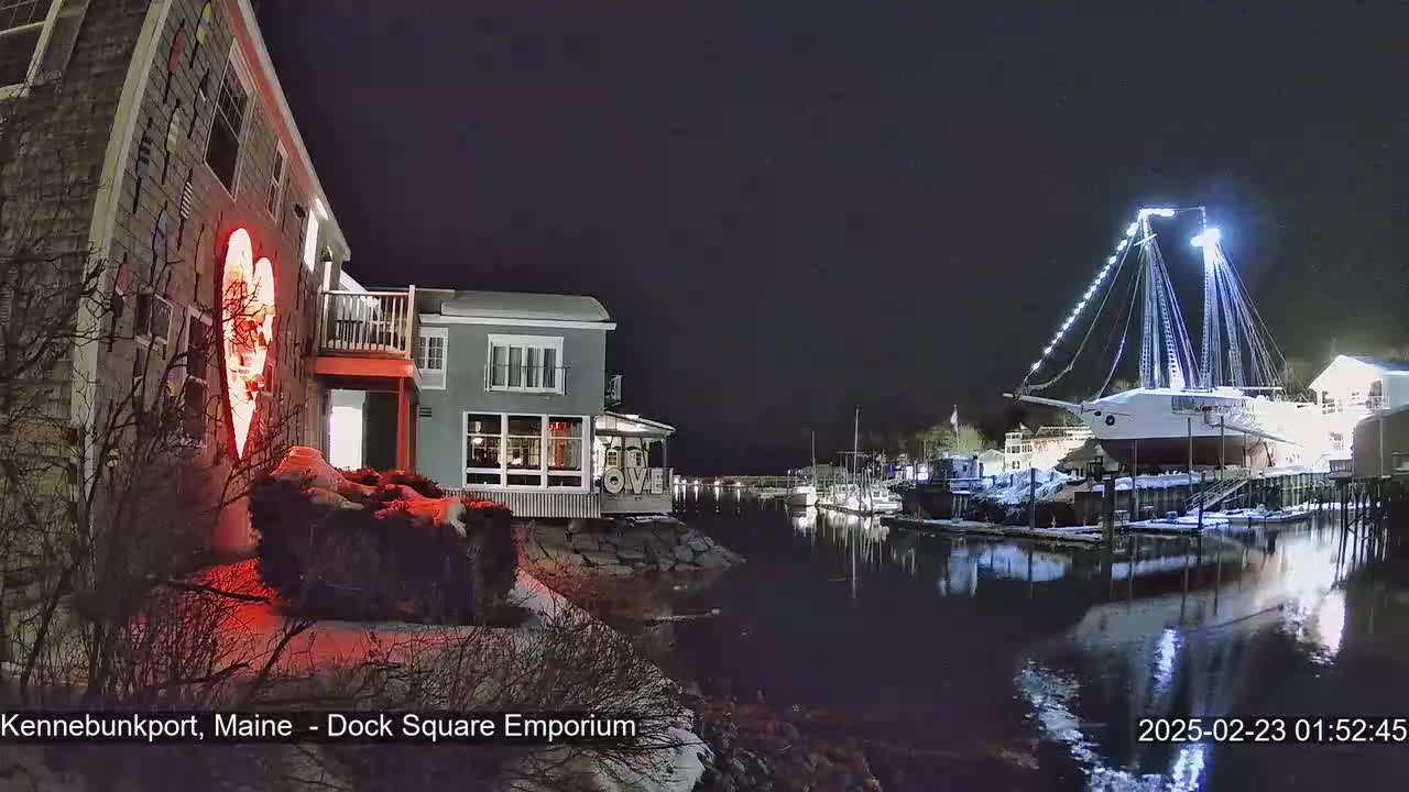 A nighttime scene shows a waterfront with a large illuminated sailboat in dry dock, buildings with lights on, and a red heart-shaped light display on the side of one building, all lightly dusted with snow.