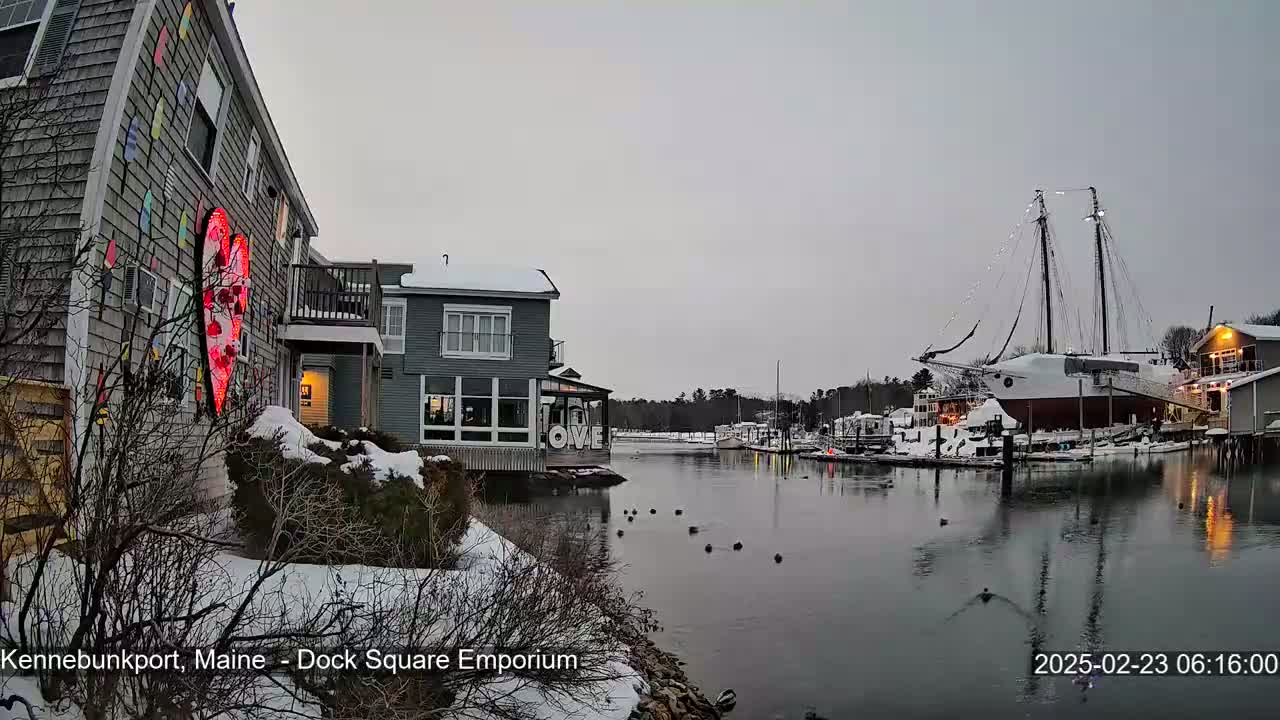 A snow-covered waterfront scene shows a building with a large illuminated heart on its side, other buildings, a large sailboat docked, and calm water with some birds.