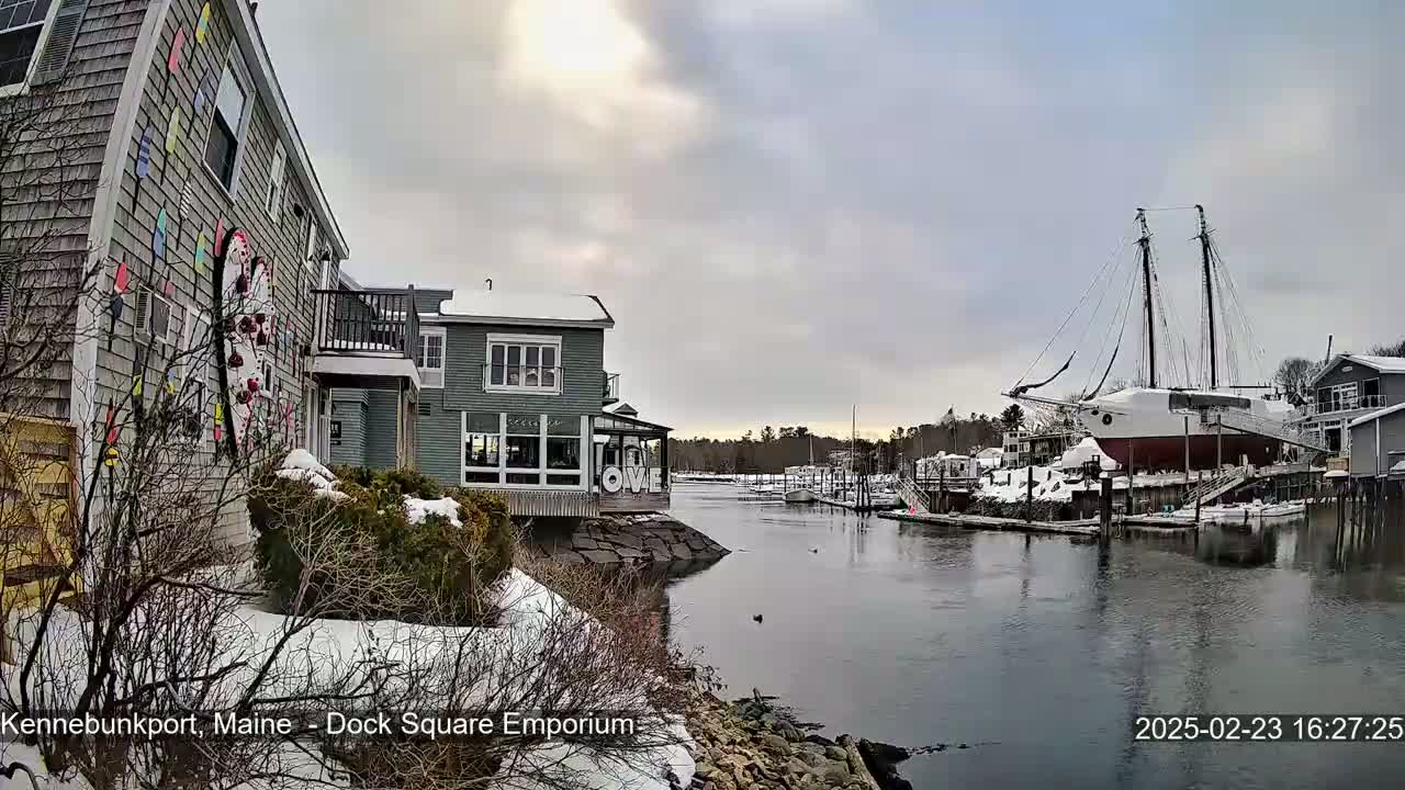 A snow-covered waterfront scene shows a harbor with docked boats, including a large sailing ship, and buildings with a colorful mural on one.