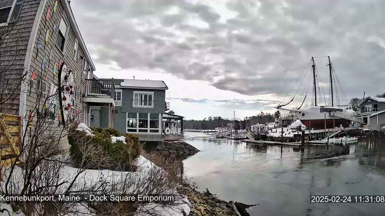 A snow-dusted waterfront scene shows buildings, a large sailboat, and a calm river under a cloudy sky.