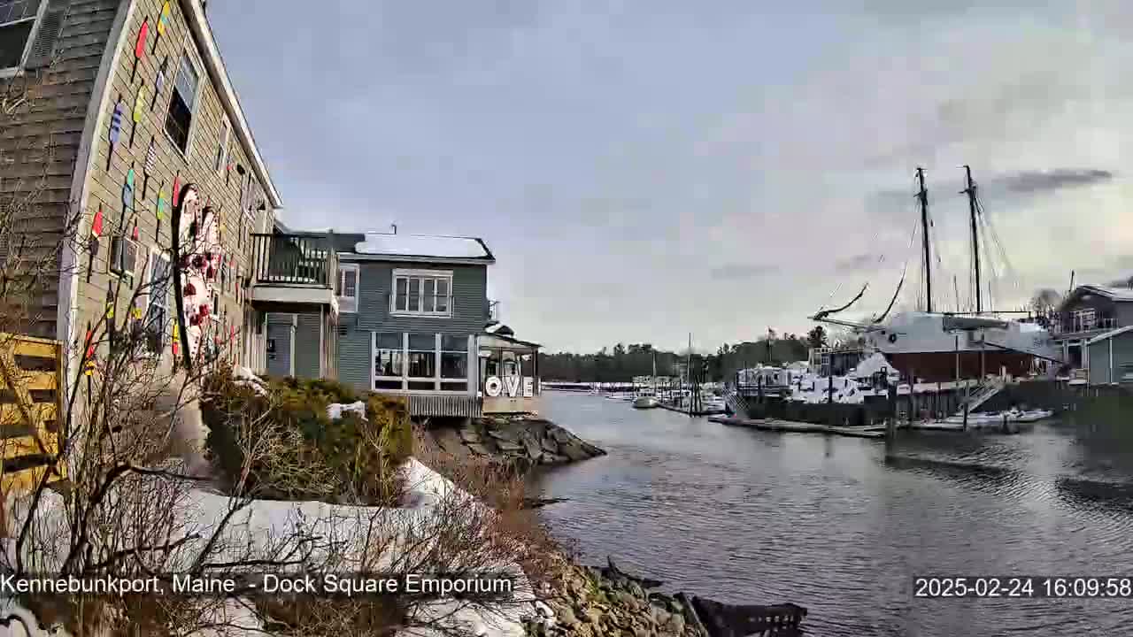 A snow-dusted waterfront scene shows a harbor with docked boats, including a large sailing vessel, next to buildings with some snow on the ground under a cloudy sky.