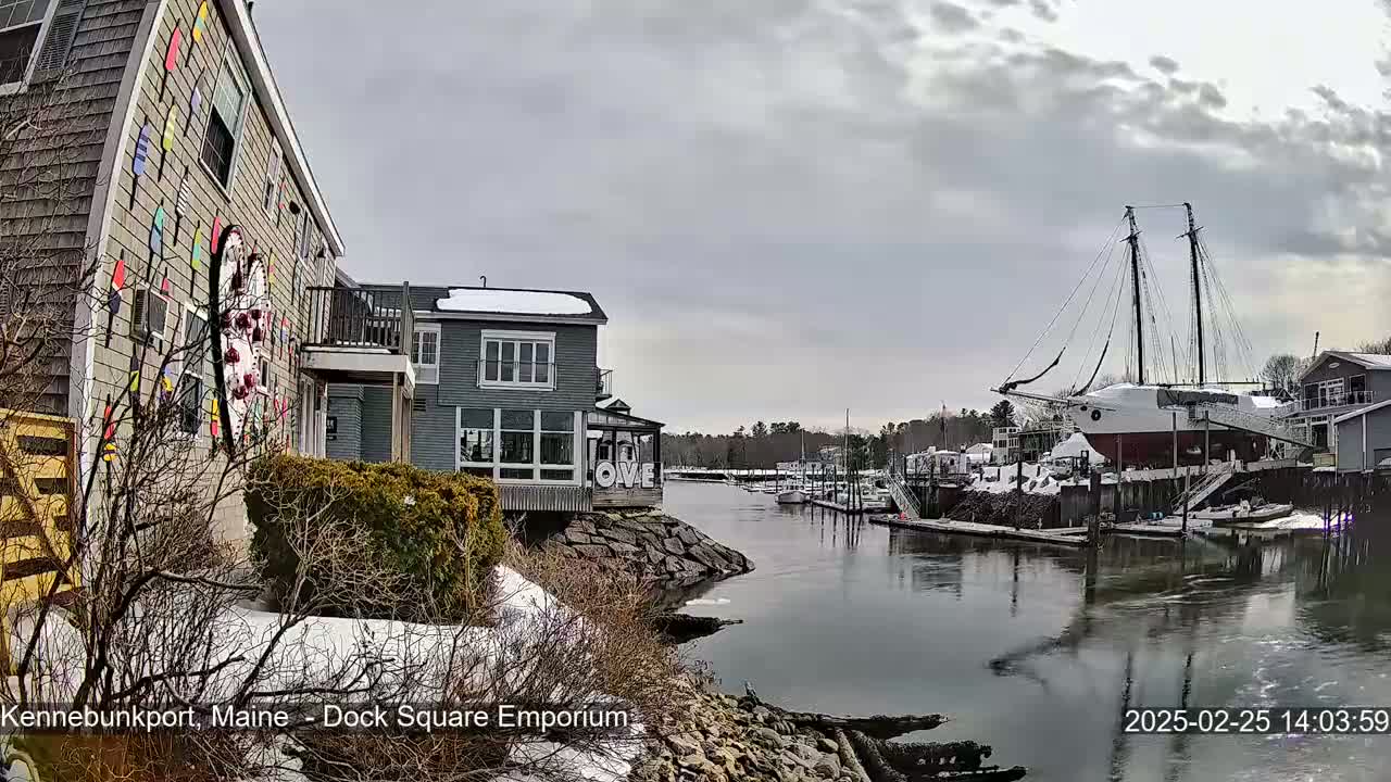 A waterfront scene on an overcast day shows a canal with docked boats, buildings with snow on their roofs, and a large sailboat on land.