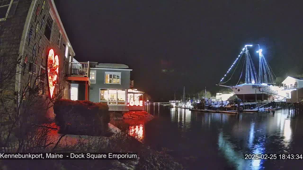 A nighttime waterfront scene shows a building with a large illuminated heart on its exterior, a docked sailboat decorated with lights, and calm water reflecting the lights.