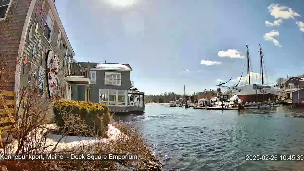 A waterfront view on a sunny day shows a building with whimsical artwork, other buildings, and a large sailboat docked in a calm waterway.