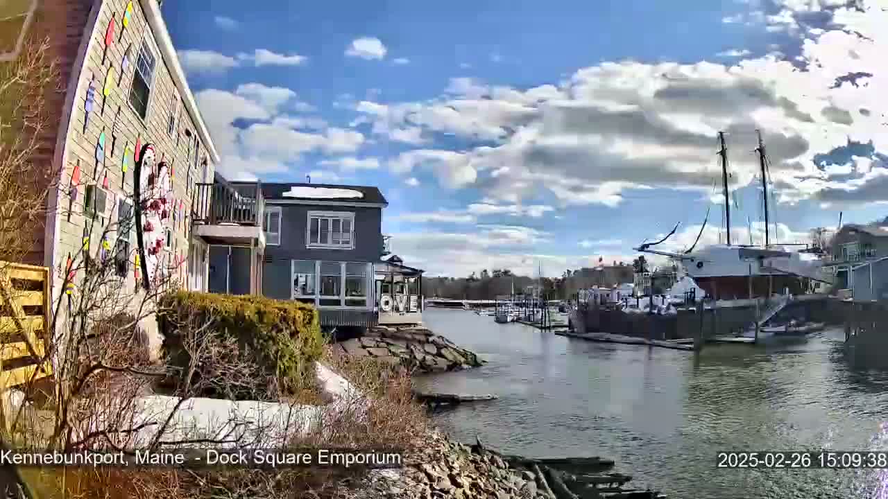 A partly sunny day shows a waterfront view of buildings, a docked sailboat, and a small amount of snow on the ground.