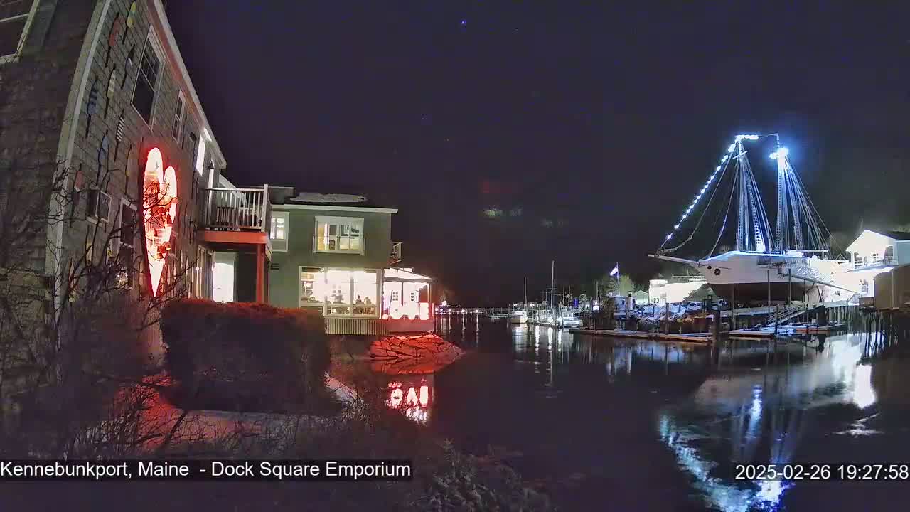 A nighttime waterfront scene shows a building with a large illuminated heart on its exterior, other buildings with lights on, a docked ship illuminated with white lights, and calm water reflecting the lights.