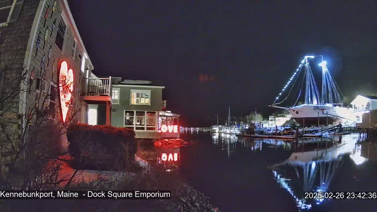 A nighttime waterfront scene shows a building with a lit-up heart on its exterior, a large sailboat illuminated with lights, and other boats docked nearby, all reflecting on the calm water under a dark sky.