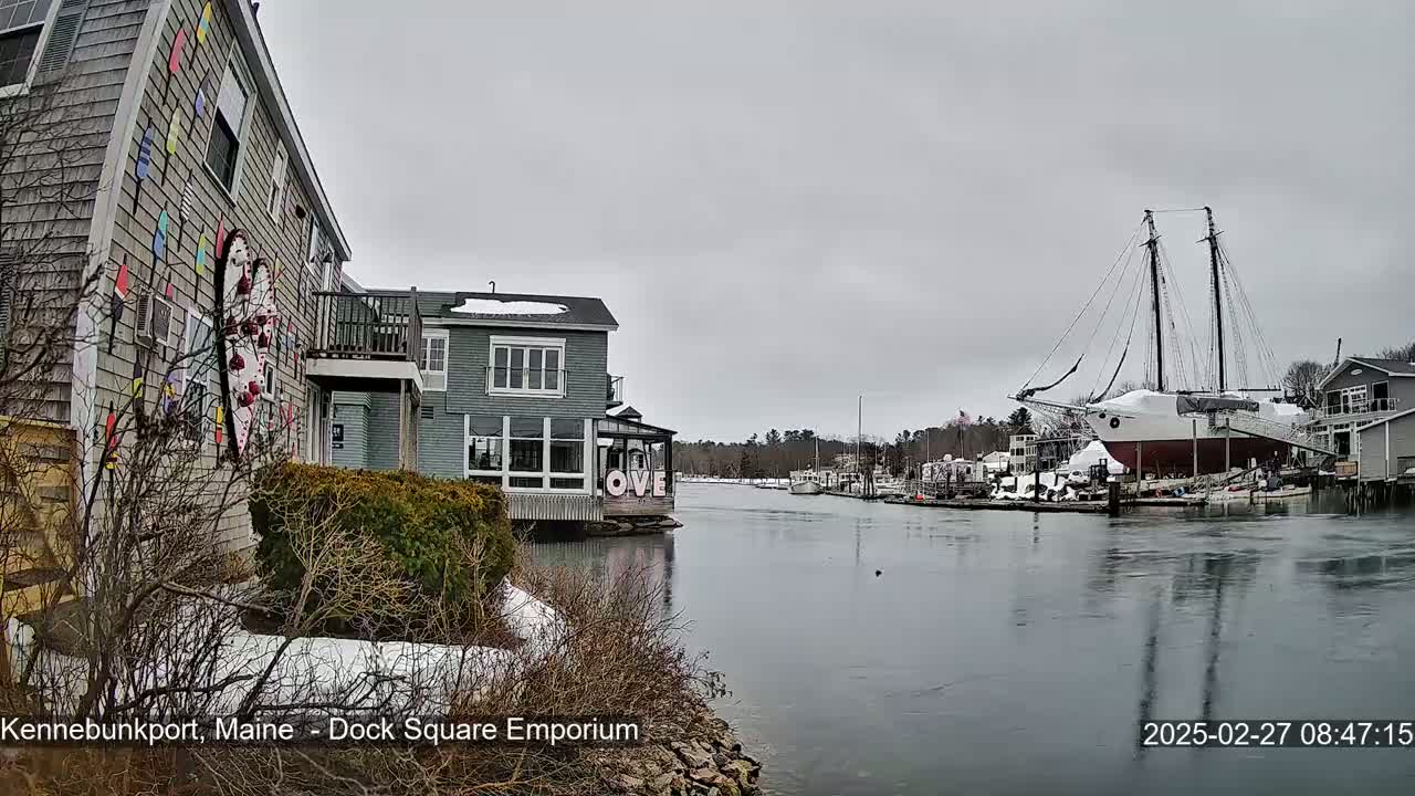 A snow-dusted waterfront scene shows a canal with docked boats, including a large sailboat, under an overcast sky.