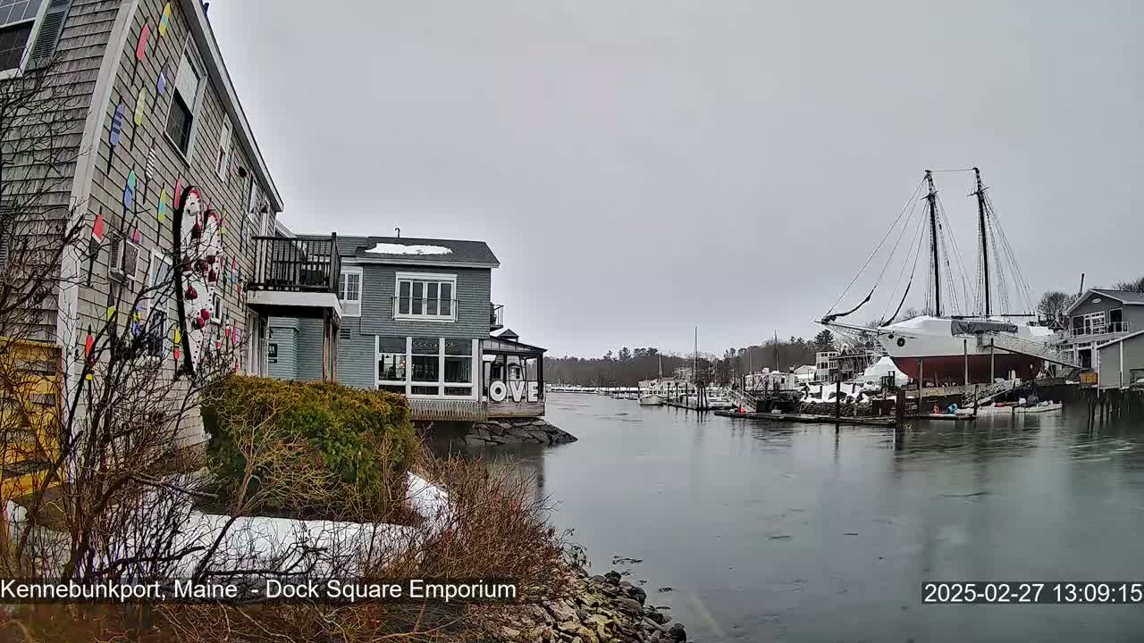A waterfront scene on an overcast day shows a gray building with colorful decorations, other buildings along a calm waterway, and a large sailboat at a dock.