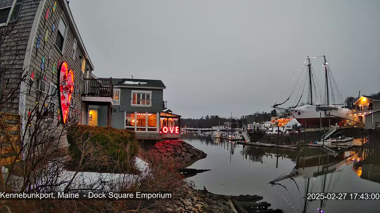 A waterfront scene at dusk shows a building with a large illuminated heart and the word "LOVE" on its exterior, a docked sailboat, and calm water reflecting the buildings and sky.