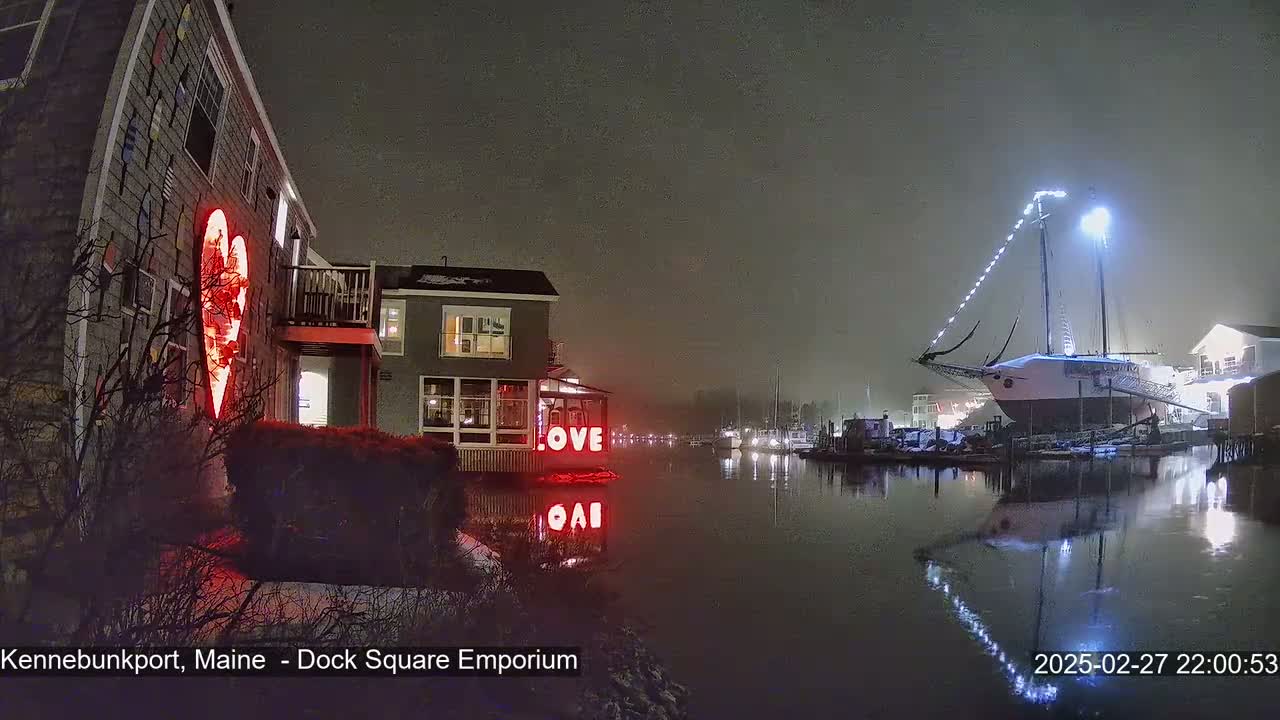 A nighttime harbor scene in foggy conditions shows a building with a large illuminated heart on its exterior, a docked sailboat with lights, and the word "LOVE" in lights on a building by the water.