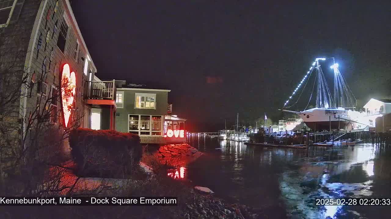 A nighttime harbor scene shows a building with a large illuminated heart on its side, a nearby building with illuminated windows, and a large sailboat lit with white lights docked in the water, all with a light dusting of snow on the ground.