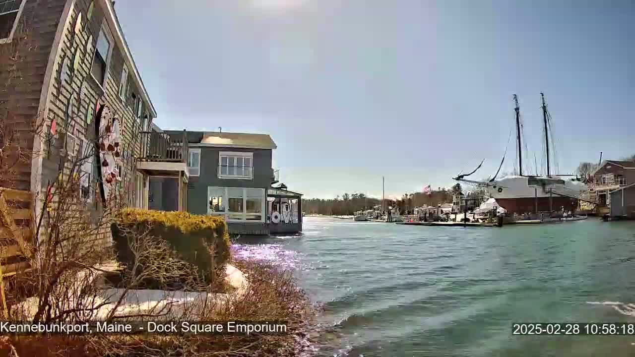 A large sailing ship is docked in a harbor on a sunny day, with buildings and other boats visible in the background.