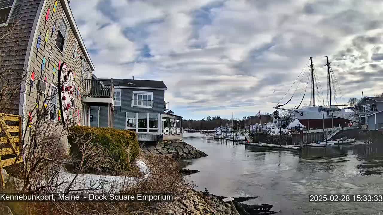 A waterfront scene on a cloudy day shows a building with colorful nautical decorations, other buildings, a docked sailboat, and a calm river with patches of snow and ice visible on the shore.