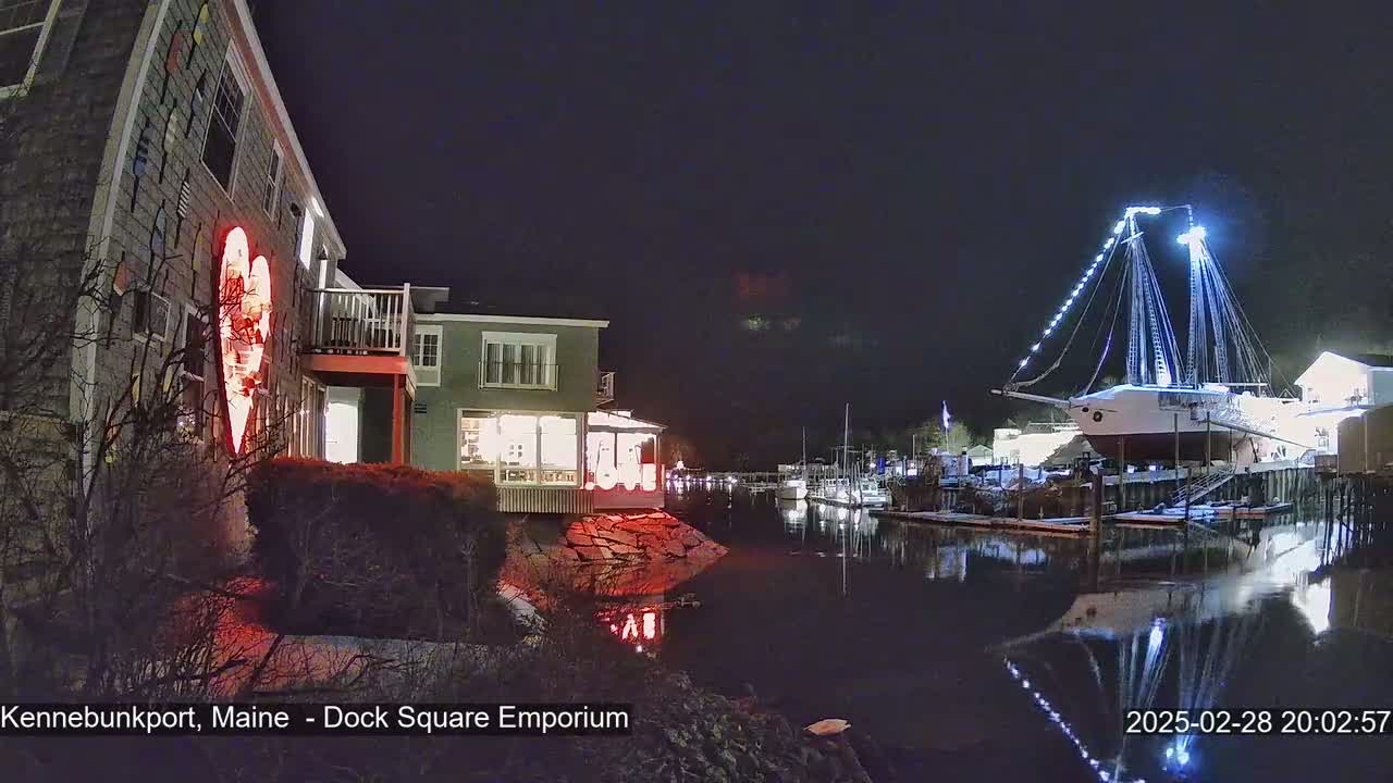 A nighttime scene shows a waterfront with a building illuminated by a large red heart, a docked ship decorated with lights, and calm water reflecting the lights.
