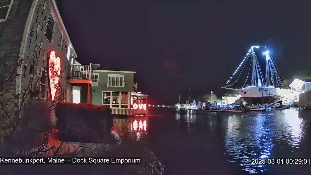 A nighttime waterfront scene shows a building with a large illuminated heart on its side, another building with the word "LOVE" displayed, and a large sailboat docked at the water's edge.