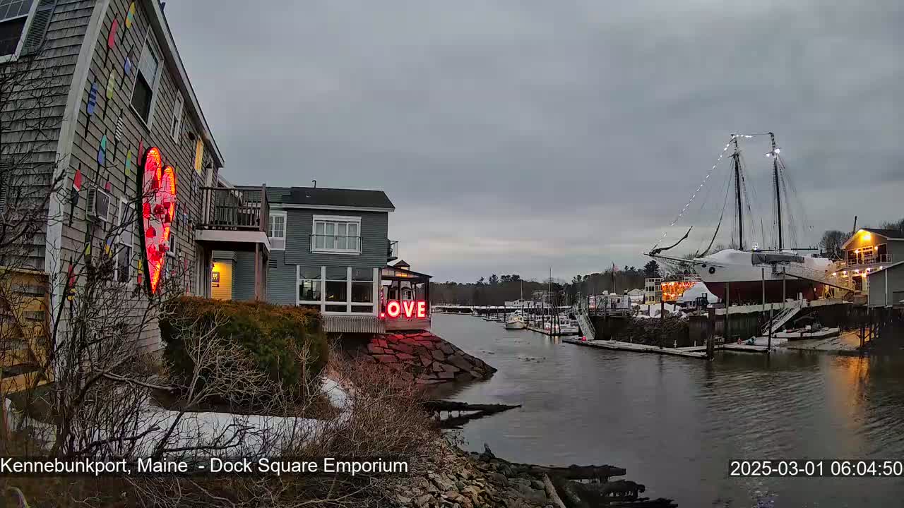 A waterfront scene under an overcast sky shows a building with a large illuminated heart and the word "LOVE" on another building, with a docked sailboat in the background, and some snow on the ground.