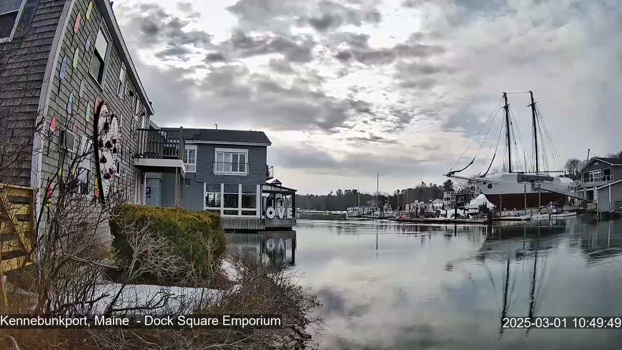 A large sailboat is docked in a calm harbor next to buildings under a cloudy sky with patches of sunlight, and there is some snow on the ground near the buildings.