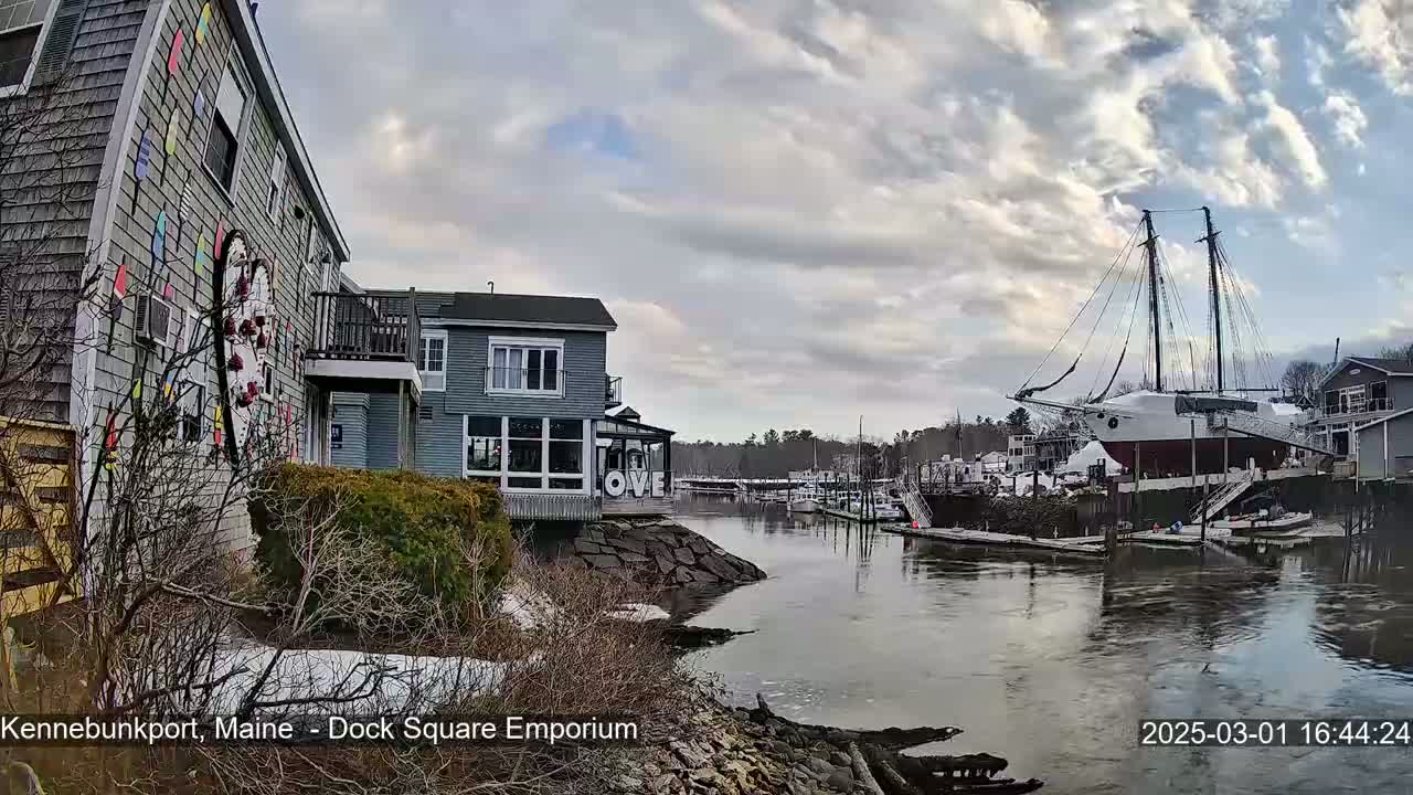 A calm waterway with docked boats and buildings is visible, under a partly cloudy sky; some snow is on the ground near the buildings.