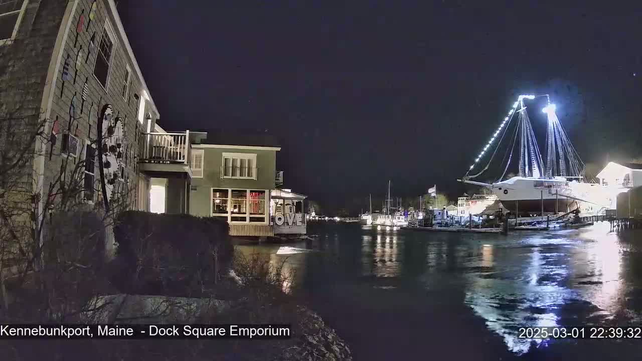 A large, brightly lit ship is docked at night next to a calm body of water, with smaller boats and buildings visible in the background.