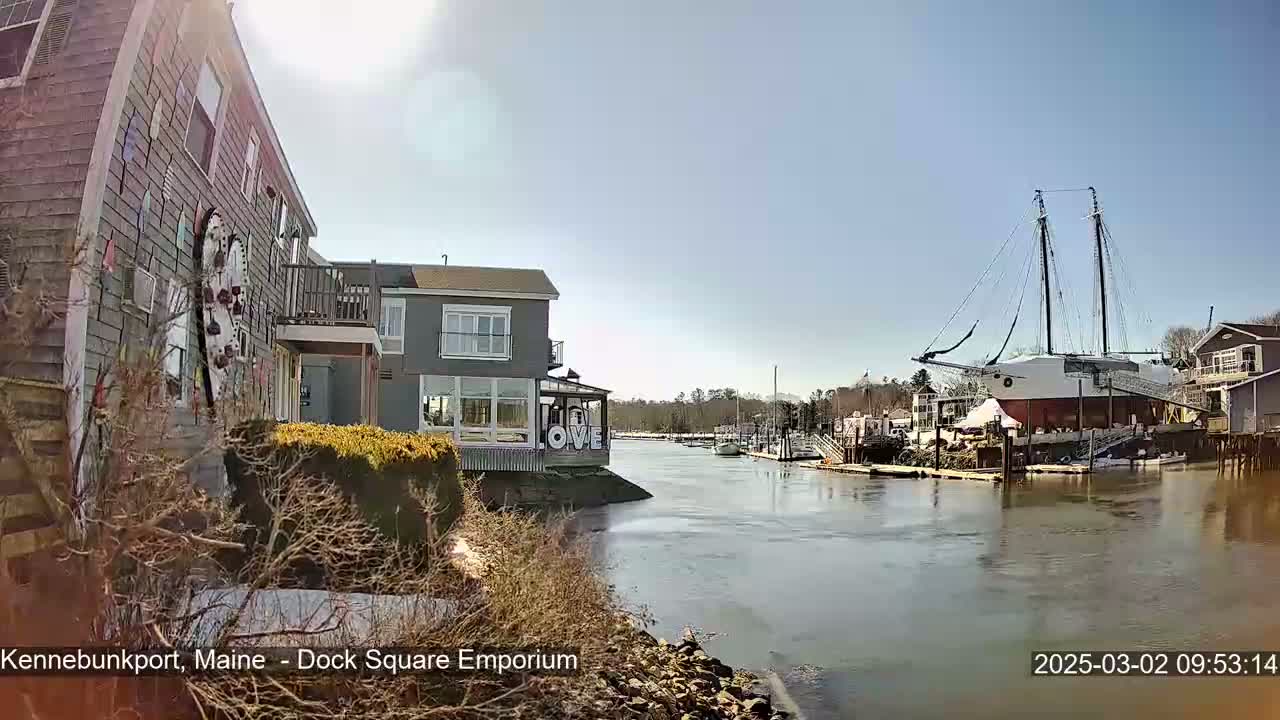 A waterfront view on a sunny day shows a gray building decorated with colorful art, a docked sailing ship, and a calm river with other boats in the distance.