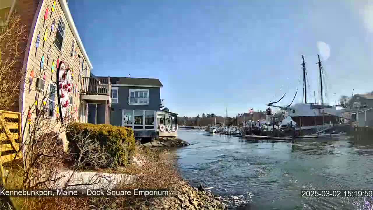 A waterfront view on a sunny day shows a building decorated with colorful nautical flags, other buildings, boats docked at a marina, and a calm waterway.