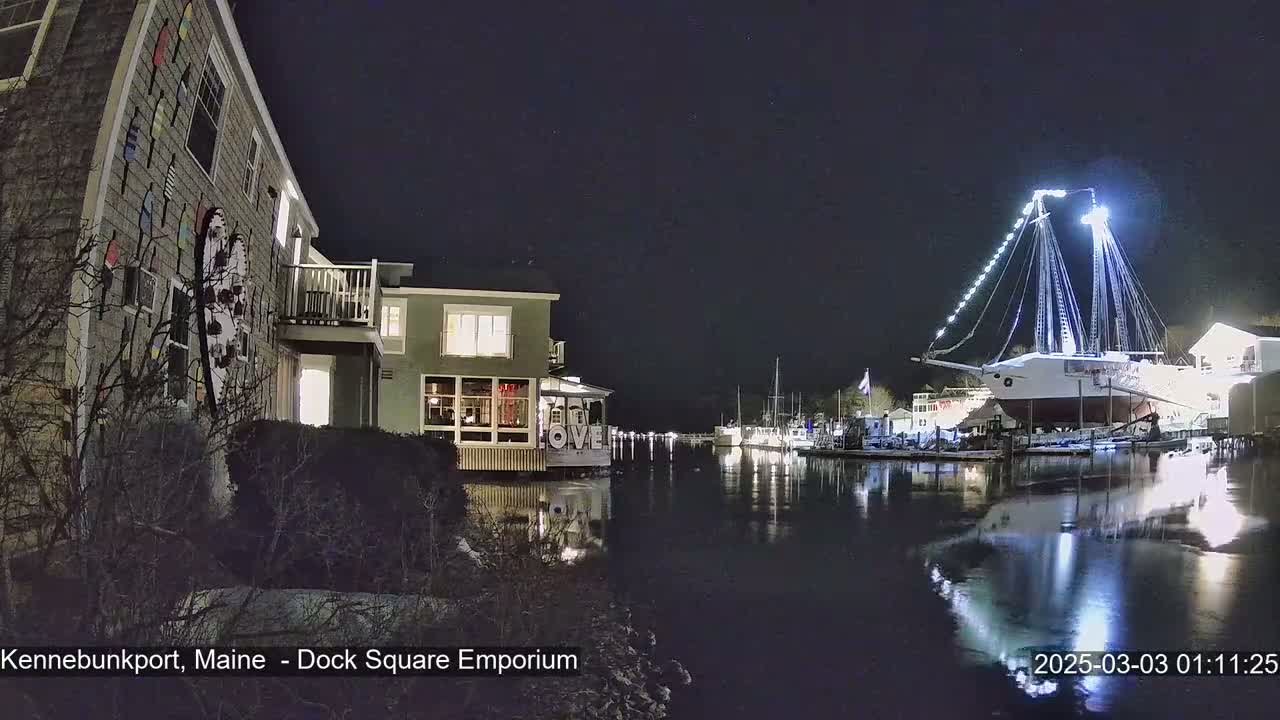 A large, illuminated sailboat is docked at a waterfront at night, near a building decorated with colorful nautical-themed items.