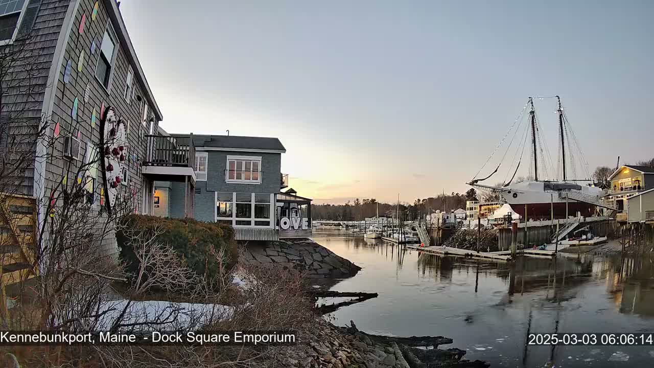 A waterfront scene at sunset shows a calm river with docked boats, including a large sailing ship, next to buildings with snow on the ground.