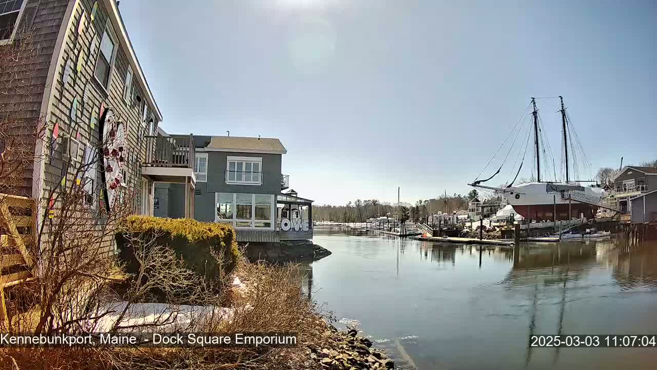 A large sailing ship rests in a calm harbor on a sunny day, next to a colorful building and other waterfront structures.