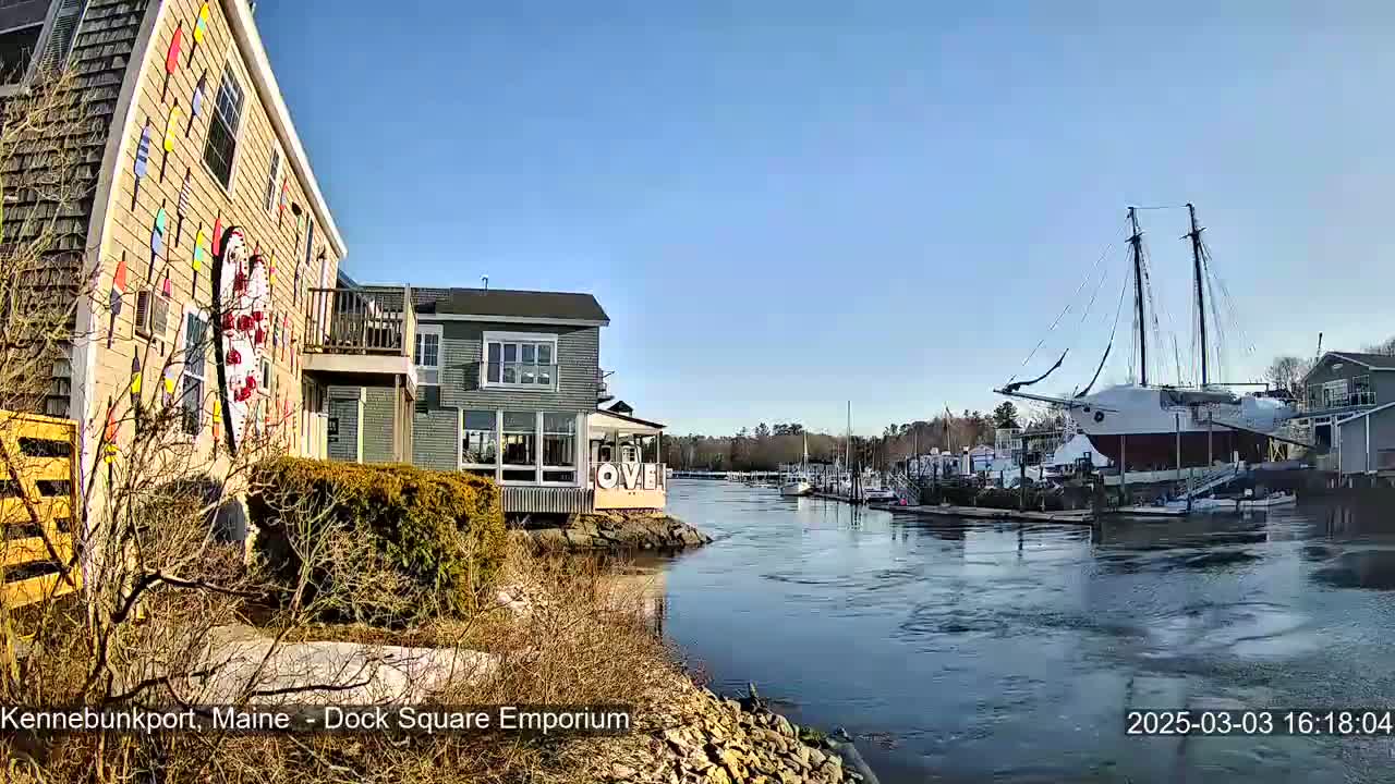 A waterfront scene on a sunny day shows a building with colorful nautical decorations next to a calm river with a large sailing ship docked nearby and other smaller boats.