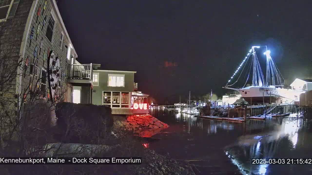 A nighttime view of a waterfront scene showing a building with colorful nautical decor, a docked illuminated sailboat, and a calm body of water with some snow on the ground.