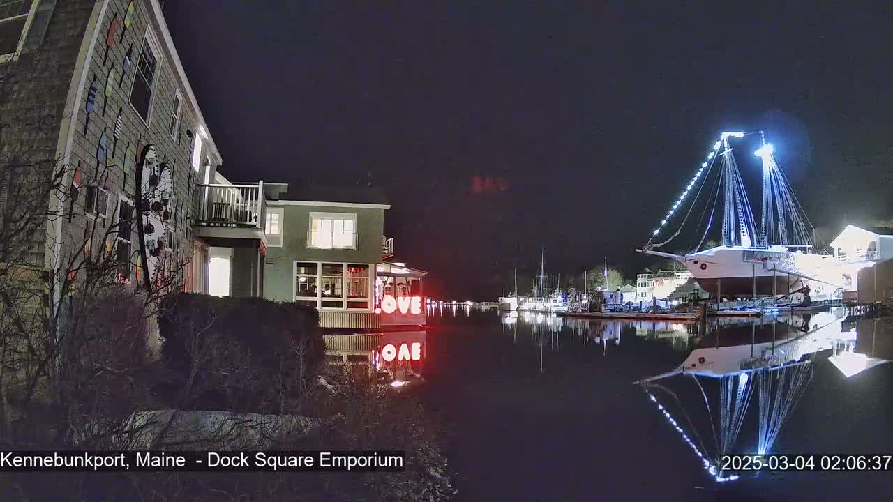 A nighttime view of a waterfront scene showing a large illuminated sailboat docked next to other boats and buildings, with the word "LOVE" in lights near the water's edge.
