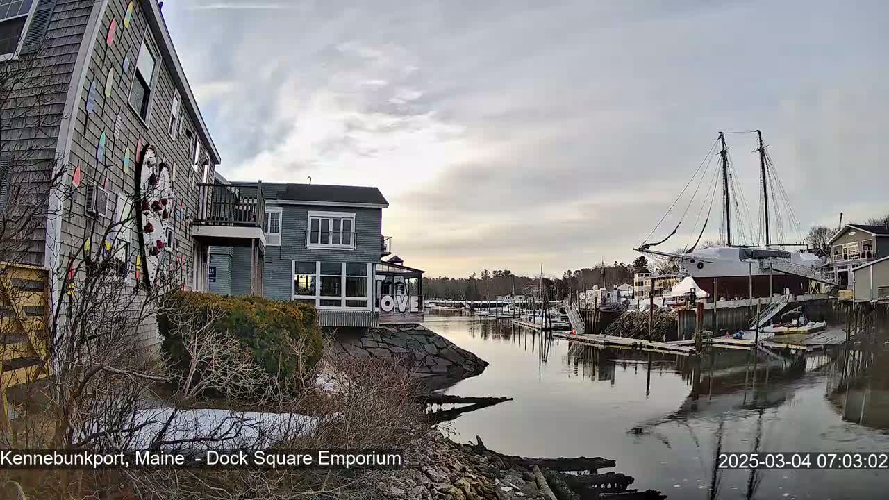 A large sailboat is docked in a calm harbor next to waterfront buildings under a cloudy sky, with some snow visible on the ground.