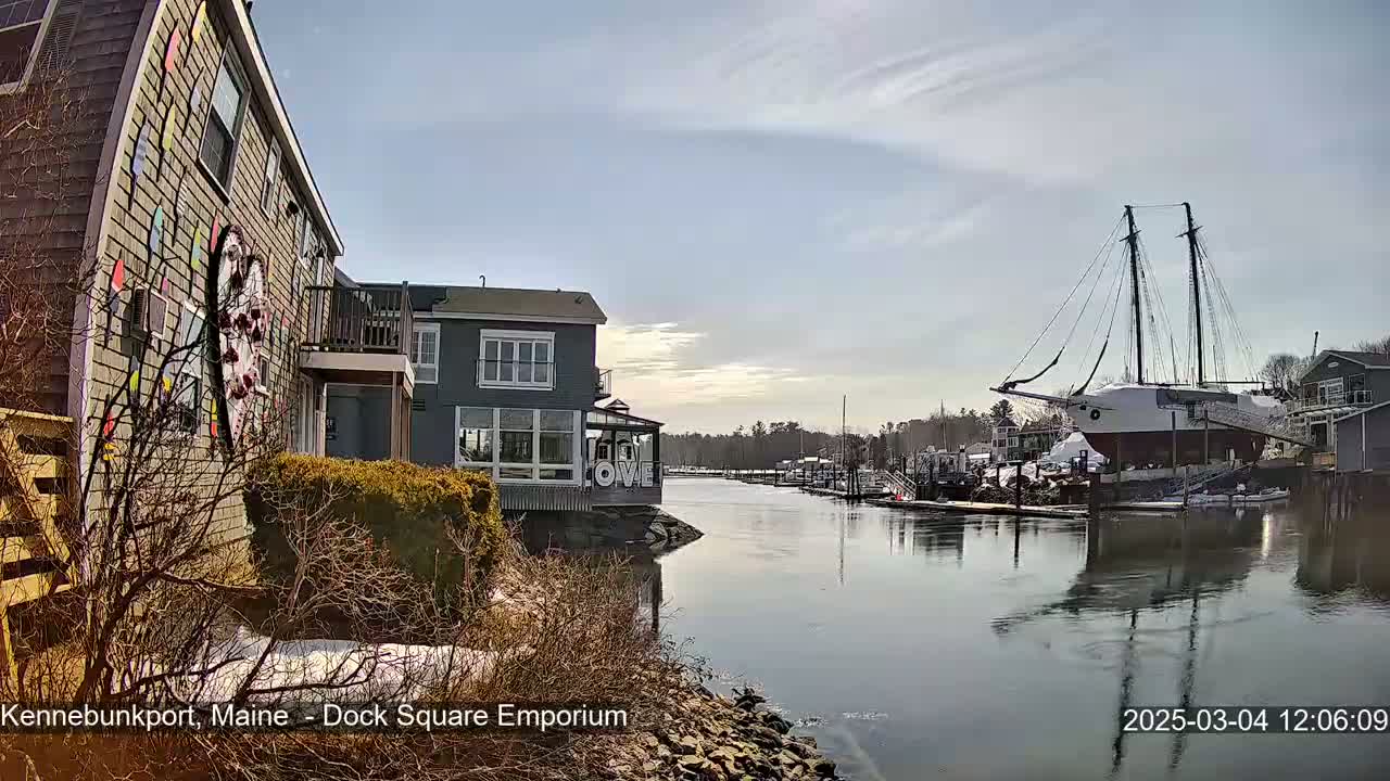 A partly sunny day reveals a waterfront scene with a large sailboat docked near colorful buildings and a calm waterway with patches of snow on the ground.