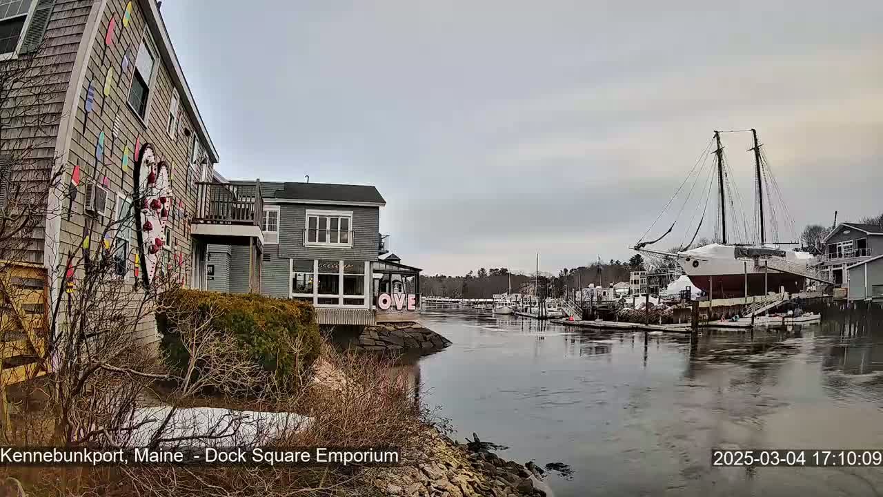 A calm, overcast day reveals a waterfront scene with buildings, a large sailboat docked, and a small body of water.