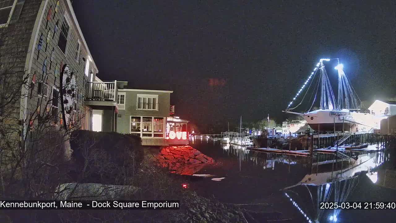 A large, illuminated sailboat rests in a harbor at night, next to a building decorated with colorful nautical designs.