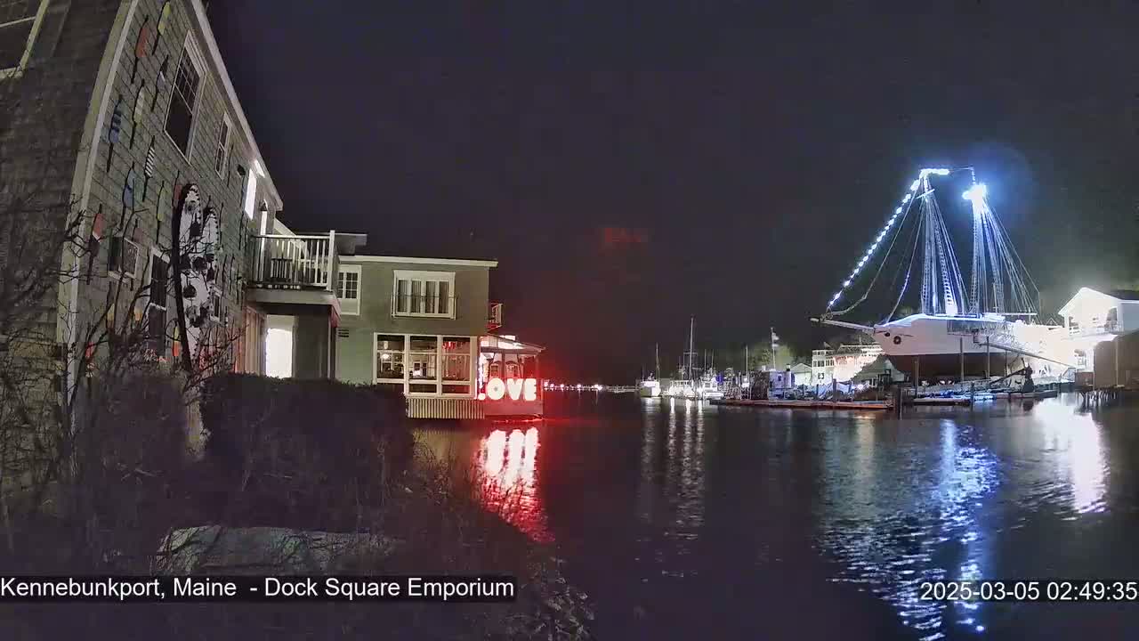 A large, illuminated sailboat is moored in a calm harbor at night, near a building with illuminated "LOVE" letters and another building decorated with nautical-themed artwork.