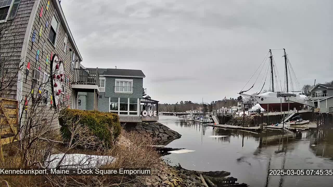 A gray day shows a waterfront scene with buildings, a large sailboat on land, and a calm river with patches of ice.