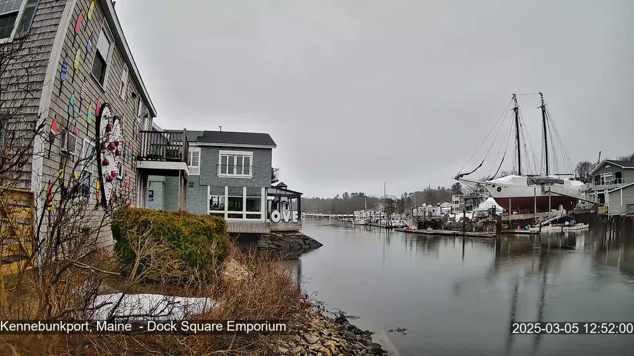 A gray, overcast day reveals a waterfront scene with a building adorned with colorful nautical decorations, other buildings, a large sailing ship on land, and a calm waterway with docked boats.