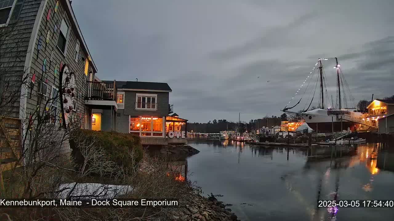 A waterfront scene at twilight shows a calm harbor with a large sailboat, several buildings with warm interior lighting, and bare winter trees.