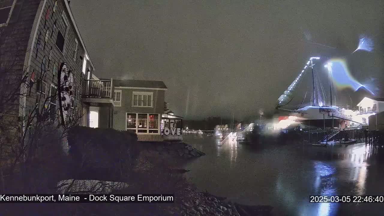 A nighttime waterfront scene shows a docked sailboat illuminated by lights, with buildings and dark vegetation visible along the shore under a dark, possibly snowy sky.