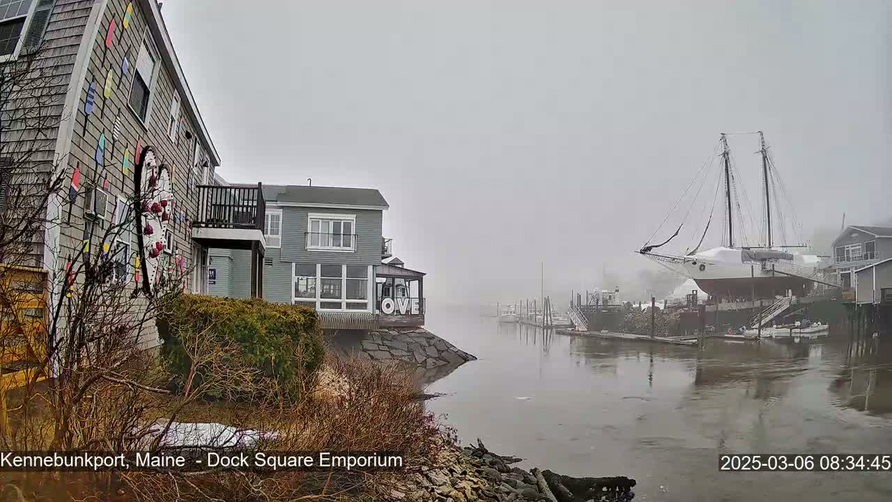 A foggy waterfront scene shows a gray building decorated with colorful oars and a large sailboat docked nearby.